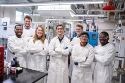A group of smiling people in white lab coats and safety glasses pose with arms crossed in an engineering lab.